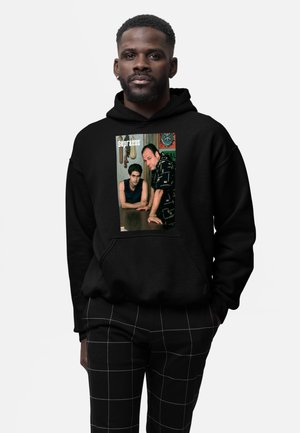 Homme portant un sweat à capuche noir avec un graphisme de deux hommes s'appuyant sur une table et le texte "Sopranos" sur le devant, assorti à un pantalon noir à carreaux.