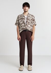 Camicia a maniche corte con colletto aperto di colore bianco con stampa di foglie marroni; abbinata a pantaloni marroni scuri e scarpe slip-on beige.