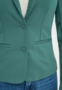 Veste teal avec une coupe ajustée, deux boutons, revers crantés et poches avant, associée à un haut crème texturé et un jean bleu.