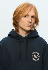 Navy blue hoodie made of soft fabric, featuring a circular logo in cream with text "WOOD WOOD COPENHAGEN 2002" on the left chest.