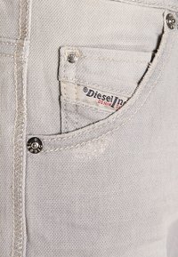 Ljusgrå denimjeans med en framficka som har en liten logotagg. Distressade detaljer och silverfärgade accenter är synliga.