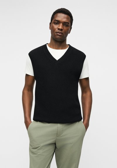 Άνδρας που φοράει μαύρο πλεκτό αμάνικο πουλόβερ με V λαιμόκοψη πάνω από λευκό μπλουζάκι και ανοιχτοπράσινο παντελόνι, με τα χέρια στις τσέπες, σε απλό φόντο.