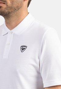 Vit pikétröja med texturerad yta, med en knappkrage och en liten svart Rossignol-logotyp på vänster bröst.