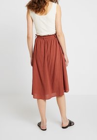 Woman wearing a sleeveless white ribbed top and a rust-colored midi skirt with gathered waist, standing barefoot on a white floor.