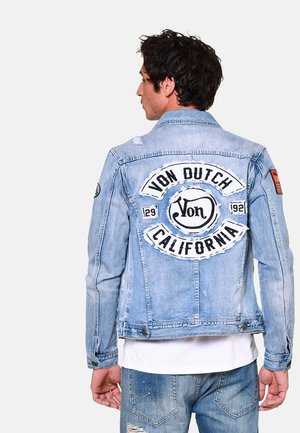 Veste en denim bleu clair avec un grand logo brodé noir et blanc à l'arrière. Détails déchirés et fermetures à boutons sont présents.
