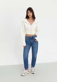 Zusammengeschnittene weiße Kapuzenjacke mit weitem Kragen und Reißverschluss, kombiniert mit hochgeschnittenen blauen Jeans und mehrfarbigen Turnschuhen. Glatte Textur.