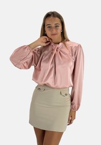 Roze satijnen blouse met lange mouwen en een strikdetail bij de hals, gecombineerd met een beige minirok met twee decoratieve knopen.