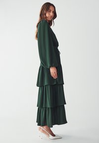Green tiered maxi dress with long sleeves and a cinched waist. Features flowing fabric and white high-heeled shoes. Textured surface.