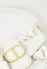 Sac à main en cuir blanc avec des accents floraux, quincaillerie dorée, un fermoir avec un logo "T" et un design élégant et rectangulaire.