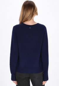 Frau mit langen hellbraunen Haaren, die einen marineblauen Pullover und schwarze Jeans trägt, von hinten vor einem weißen Hintergrund gezeigt.