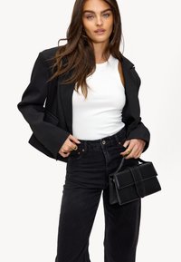 Zwarte blazer over een witte tanktop, gecombineerd met hoge zwarte jeans. Houdt een gestructureerde zwarte handtas vast met een bovenflap.