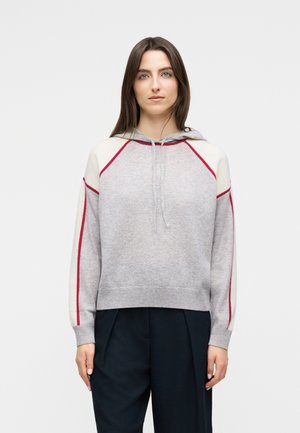 Γυναίκα που φοράει γκρι ανοιχτό φούτερ με κόκκινη επένδυση στα μανίκια και τους ώμους, συνδυασμένο με μαύρο παντελόνι, στέκεται μπροστά σε απλό φωτεινό φόντο.