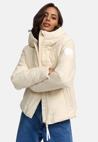 Doudoune beige avec col haut, design matelassé horizontal, poches latérales et détail logo sur la manche. Associée à un jean bleu.