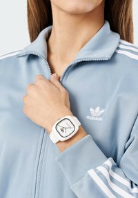 Valge ruudukujuline kell valge rihmaga ning musta ja valge näoga, kantud helesinise tõmblukuga jaki peal, millel on valged triibud ja logo.