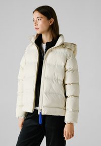 Cremfarbige Puffjacke mit abnehmbarer Kapuze, gesteppter Optik, Reißverschluss und blauen Reißverschlussanhänger, kombiniert mit einer schwarzen Zip-Jacke darunter.