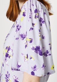 Femme portant un chemisier texturé violet clair avec un imprimé floral violet et jaune et des manches courtes amples, vue de profil.