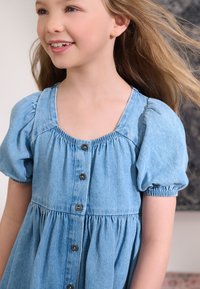 Spijkerjurk in lichtblauw, met korte pofmouwen, een vierkante halslijn en een knoopsluiting aan de voorkant met donkere knopen. Gestructureerde stof.