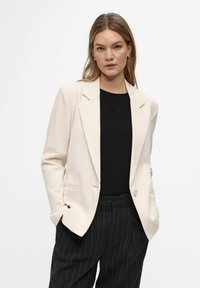 Blazer beige de tela texturizada, con solapa en pico, cierre de un solo botón y dos bolsillos frontales, combinado con pantalones de rayas diplomáticas.
