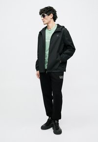 Zwarte lichtgewicht jas met capuchon, groene T-shirt eronder, zwarte trainingsbroek met logo en zwarte sneakers. Model draagt een zonnebril.