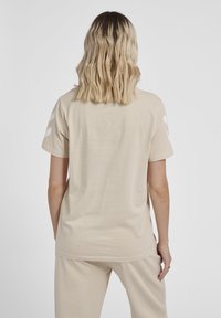 Beige bomulds T-shirt med afslappet pasform, korte ærmer og tre hvide vandrette striber på hver ærme. Glat tekstur, enkelt design.
