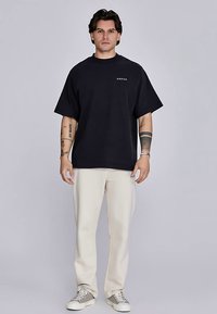 Black oversized t-shirt with a round neckline, short sleeves, and a small white logo. Paired with cream-colored tapered trousers and sneakers.
