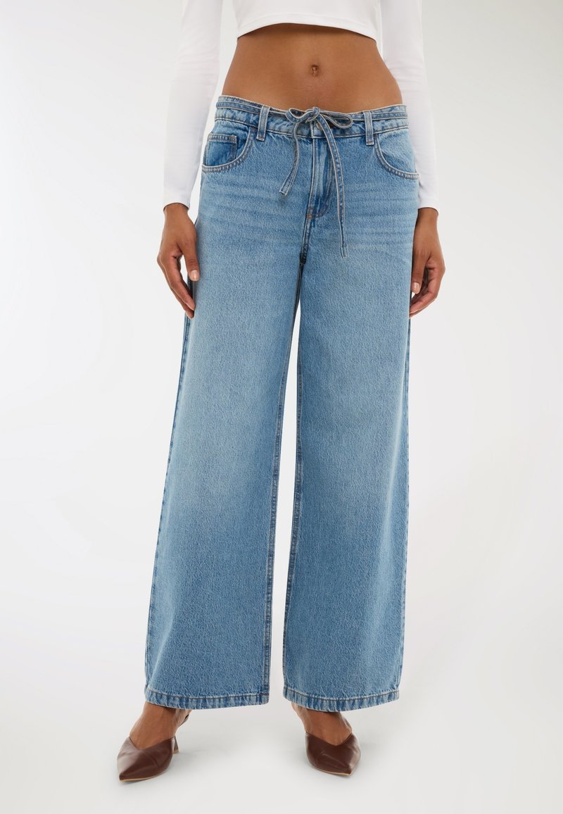 Jasnoniebieskie dżinsy o szerokich nogawkach, wykonane z denimu, z elastycznym pasem na sznurki, przednimi kieszeniami i surowymi brzegami. W połączeniu z brązowymi butami na obcasie.