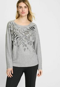 Grijze longsleeve trui met ronde halslijn en een grafische print in zwart. Zachte textuur met een ontspannen pasvorm, gecombineerd met donkere broeken.