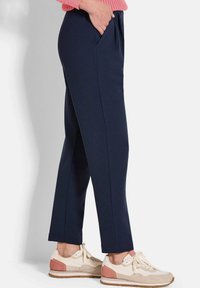 Personne portant un pantalon tapered bleu marine et des baskets crème avec des accents rose pâle, main dans la poche, sur un fond clair et uni.