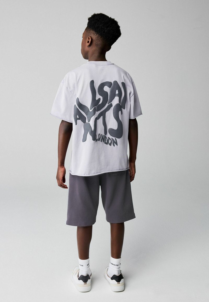 Poiss lühikeste lokkis juustega, seljas valge liialdatud suurusega T-särk, millel on seljal suur must „ALLSAINTS LONDON” kiri, hallid lühikesed püksid ja valged tossud.