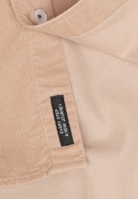 Beige corduroy stof met verticale ribbels; bevat een zwart genaaid label met witte tekst: "ELKE STAP EEN NIEUWE REIS." Inclusief een knoopsluiting.