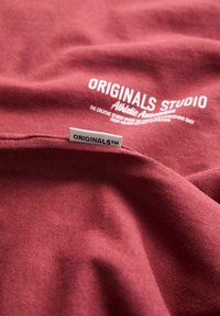 Burgundy bomuldsstof med en glat tekstur. Har hvide tryk med teksten "ORIGINALS STUDIO Athletic Assortments" og et lille mærke, der er mærket "ORIGINALS™."