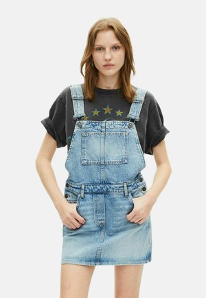 Jeans-Overall-Kleid in Hellblau mit verstellbaren Trägern, Fronttaschen und einer taillierten Taille. Getragen über einem dunklen Grafik-T-Shirt.