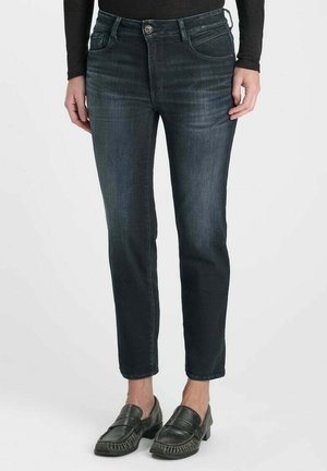 Personne portant un jean slim bleu foncé associé à des mocassins noirs et un haut noir à manches longues, debout devant un fond uni.