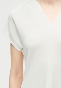 Κοντινό πλάνο ενός ατόμου που φοράει λευκό κοντομάνικο μπλουζάκι με V λαιμόκοψη και λεία υφή, σε απλό φωτεινό φόντο.