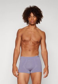 Mladic s kovrčavom kosom koji nosi prilagođene svijetloplave bokserice, stoji protiv jednostavne svijetle pozadine, okrenut prema naprijed.