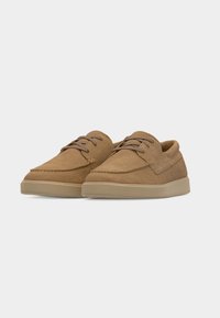 Paire de chaussures casual en daim marron clair à lacets avec semelles en caoutchouc beige sur un fond gris clair uni.