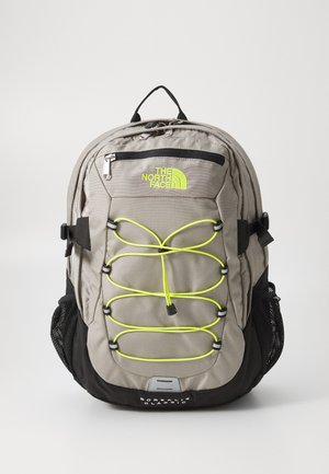 Zaino in tessuto grigio con accenti neri. Presenta una tasca frontale con zip, un design con corda bungee gialla fluorescente e tasche laterali in rete. Logo del brand visibile.