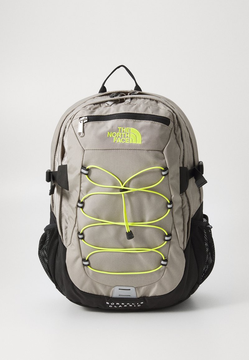 Mochila de tecido cinza com detalhes pretos. Apresenta um bolso frontal com fecho, um design de corda elástica amarelo-neon e bolsos laterais em rede. Logótipo da marca exibido.