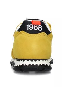 Sneaker giallo in suede con tallone nero, caratterizzato da dettagli rossi e bianchi e suola a strisce zigzag. Sul retro è visibile il numero "1968."