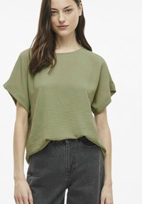 Olivinės spalvos, tekstūruota trumpo rankovės palaidinė su apvaliu iškirpimu, derinama su tamsiomis, aukštai įstumtomis džinsais. Nėra matomų raštų ar akcentų.