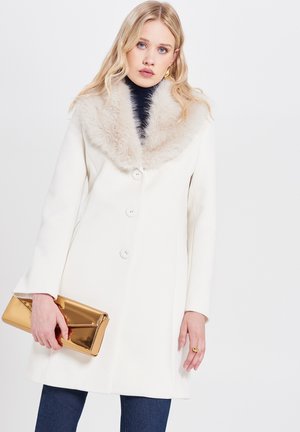 Cappotto bianco con colletto di pelliccia, dotato di tre bottoni, tasche laterali e una texture liscia. La modella tiene in mano una pochette dorata.