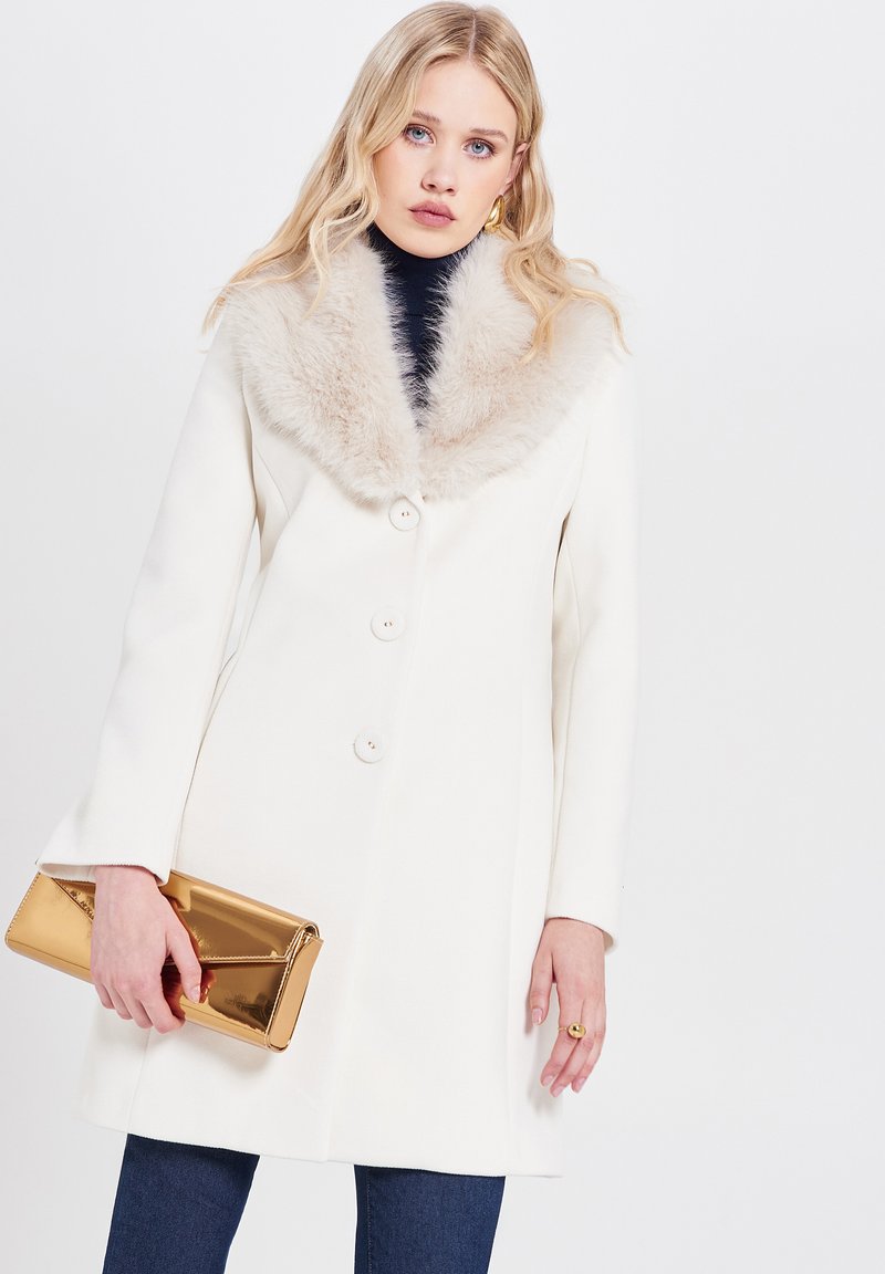 Cappotto bianco con colletto di pelliccia, dotato di tre bottoni, tasche laterali e una texture liscia. La modella tiene in mano una pochette dorata.