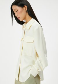 Blusa gialla chiara in tessuto morbido, con colletto a punta, tasche frontali e patta con bottoni. Vestibilità ampia con maniche a polsino.