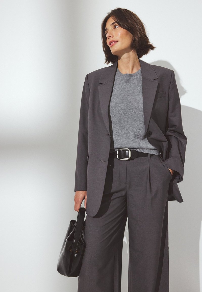 Pilkas švarkas su pilku megztiniu, derinamas su platėjančiomis pilkomis kelnėmis. Juodas diržas ir rankinė; lygus audinys su struktūrizuotu dizainu.