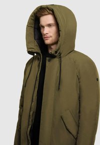 Olivza zaļa polsterēta jaka ar lielu kapuci, priekšējo rāvējslēdzēju un auklām. Iespļaujas sānu kabata un melni akcenti uz kapuces.