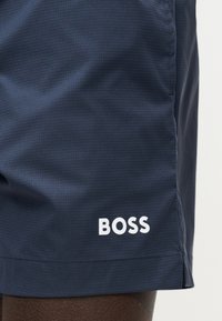 Σκούρα ναυτικά υφασμάτινα σορτς με λευκό λογότυπο «BOSS» στο κάτω στρίφωμα πάνω από το γόνατο, σε σκουρόχρωμο τόνο δέρματος.