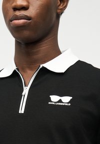 Κοντινό πλάνο ενός ατόμου που φοράει μαύρο πόλο πουκάμισο με λευκό γιακά και μερικό φερμουάρ, με λευκό λογότυπο Karl Lagerfeld με γυαλιά ηλίου.