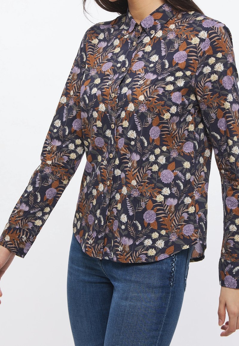 Femme portant une chemise boutonnée avec un fond sombre et un motif floral en violet, blanc et marron, assortie à un jean bleu.