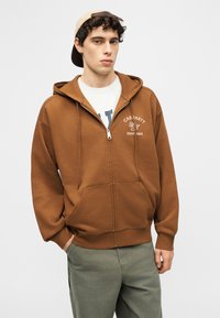 Mladý muž v hnedej mikine na zips s textom na hrudi, v bielom tričku, zelených nohaviciach a béžovej šiltovke nasadenej dozadu na jednoduchom pozadí.