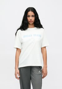 Hosszú fekete hajú nő fehér Jordan Brand pólóban és szürke sportnadrágban áll egy sima fehér háttér előtt.